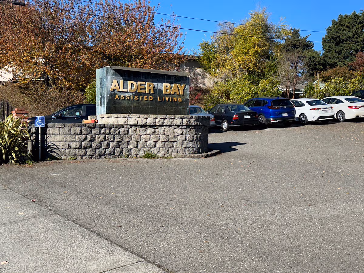 Alder Bay Assisted Living sign sits on a stone pedestal beside a row of parked cars.