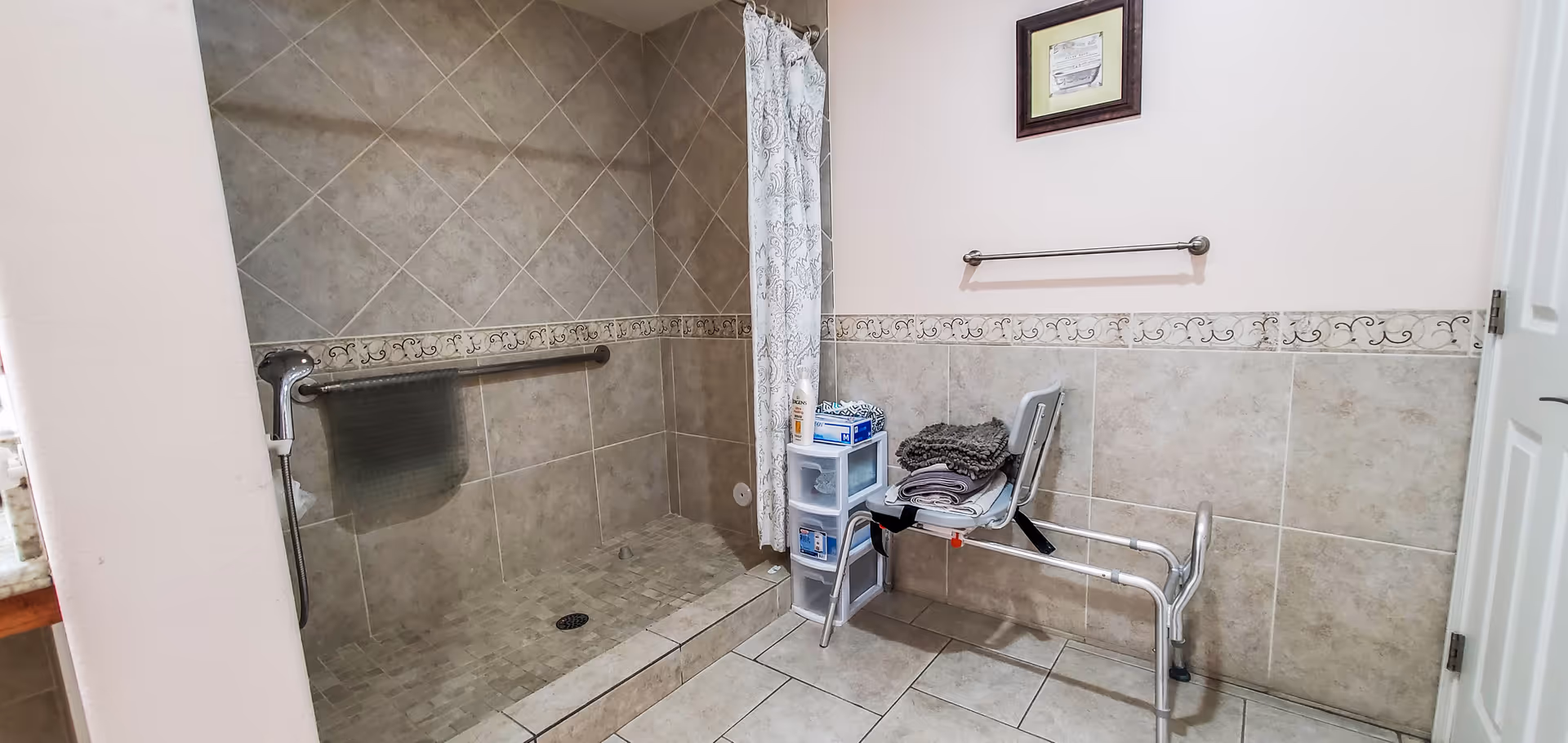 Accessible bathroom with a tiled walk-in shower, shower chair, grab bars, and a storage cart.
