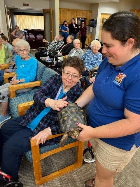 A group of elderly residents seated in a common room at a senior living facility. One woman is touching a tortoise held by a staff member wearing a blue shirt. Other residents and staff are visible in the background, some seated and some standing near a doorway.