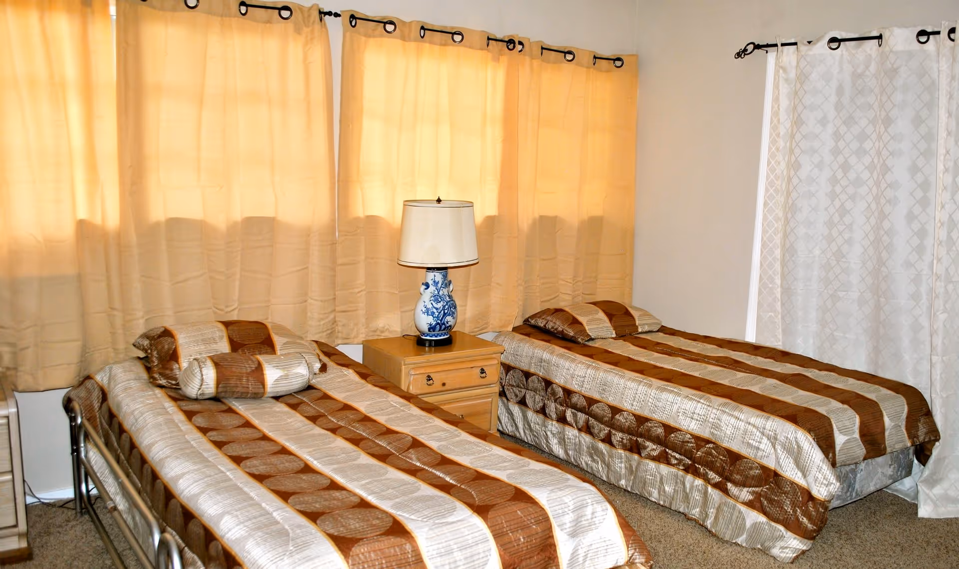A bedroom with two single beds, each covered with matching brown and beige striped bedspreads and pillows. Between the beds is a wooden nightstand with a blue and white decorative lamp. The room has beige curtains covering the windows and light-colored walls.