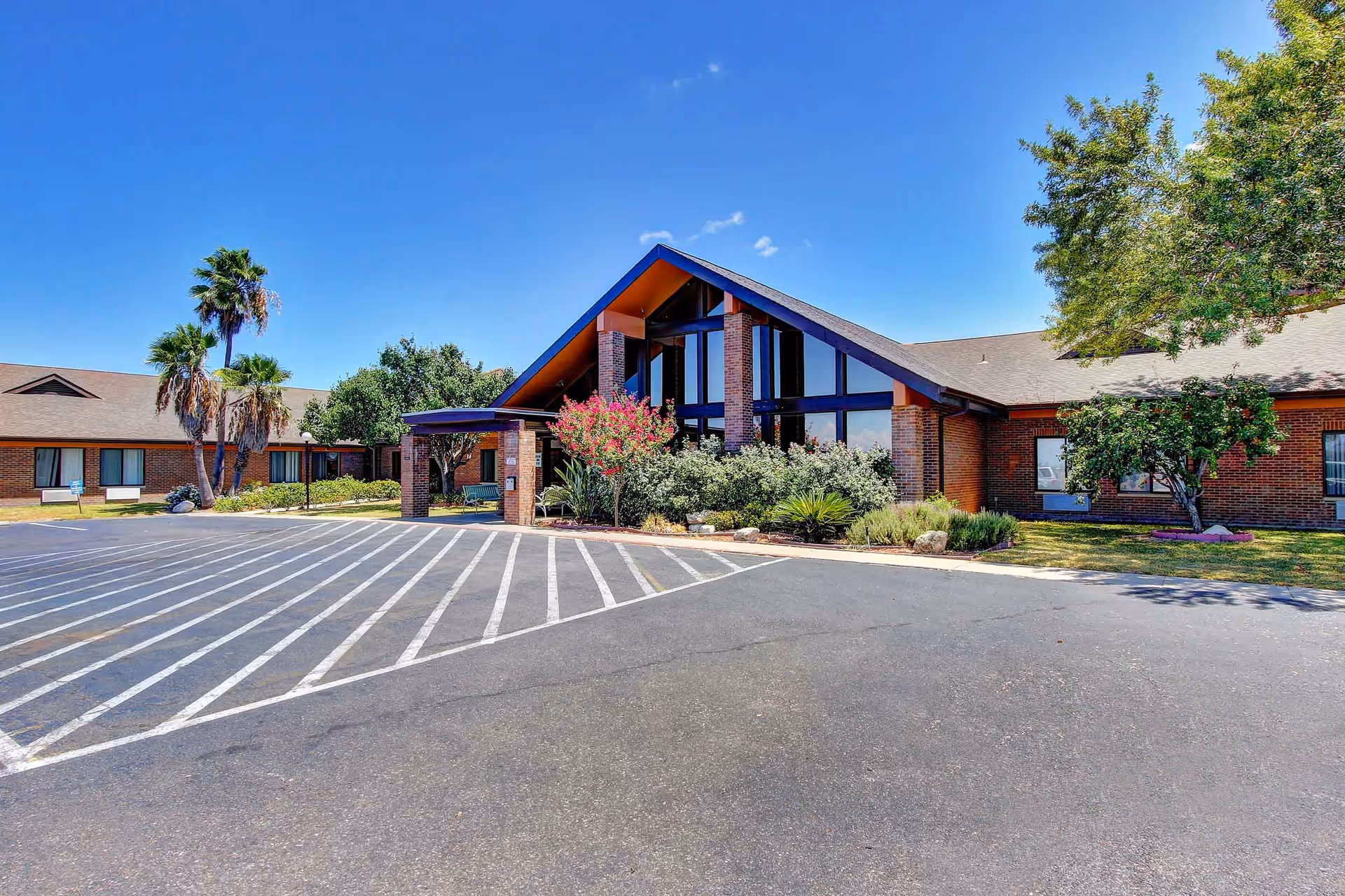 Exterior view of a single-story brick building with a large triangular glass entrance and a covered entryway. The building is surrounded by landscaping including palm trees, flowering bushes, and other greenery. The sky is clear and blue, and there is a paved parking area with white striped markings in front of the building.