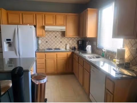 A kitchen with wooden cabinets, a white refrigerator, a gas stove with a range hood, a dishwasher, a double sink, and a window with blinds letting in natural light. The countertops are dark, and there is a trash can and a few kitchen appliances visible.