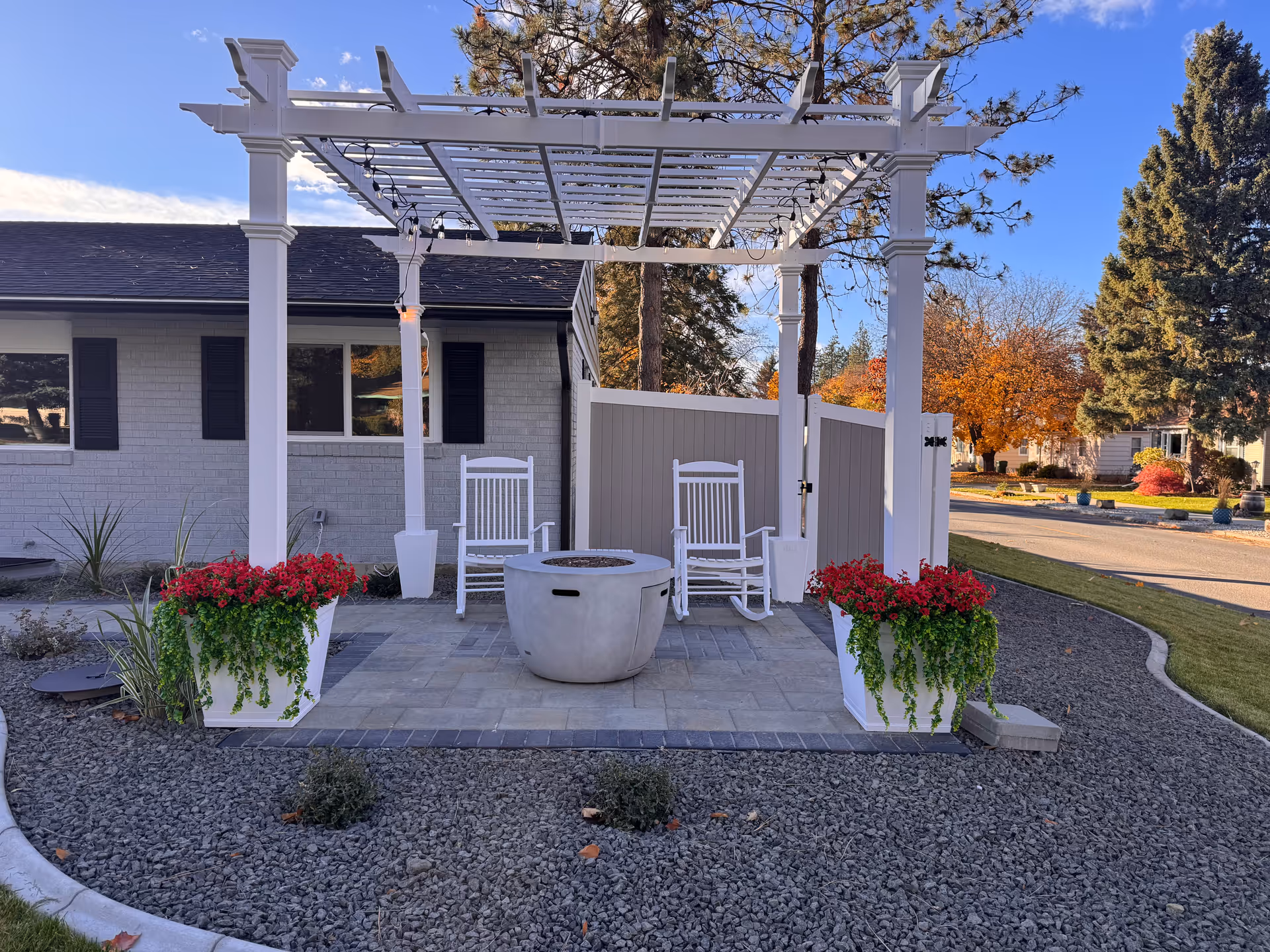 White pergola patio with rocking chairs, fire pit, and red potted flowers outside a beige building.