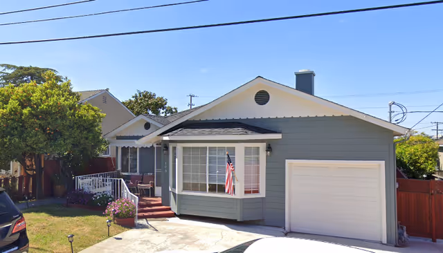 Exterior view of a single-story residential building with gray siding and white trim, featuring a garage, a bay window with an American flag, a small front porch with steps, and a well-maintained lawn with a tree and flowers under a clear blue sky.