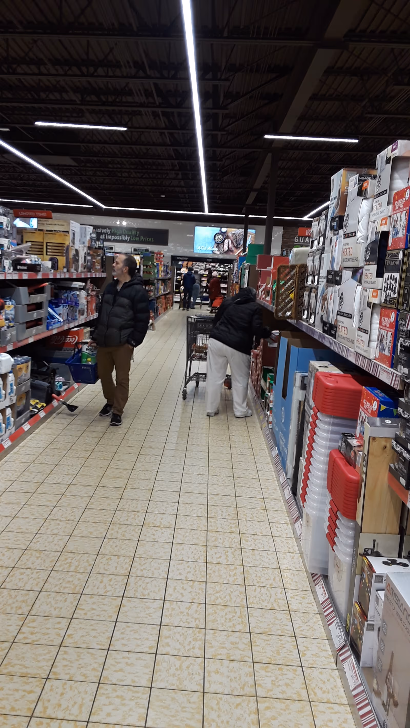 Shoppers navigate a bright supermarket aisle with tall shelves on both sides.