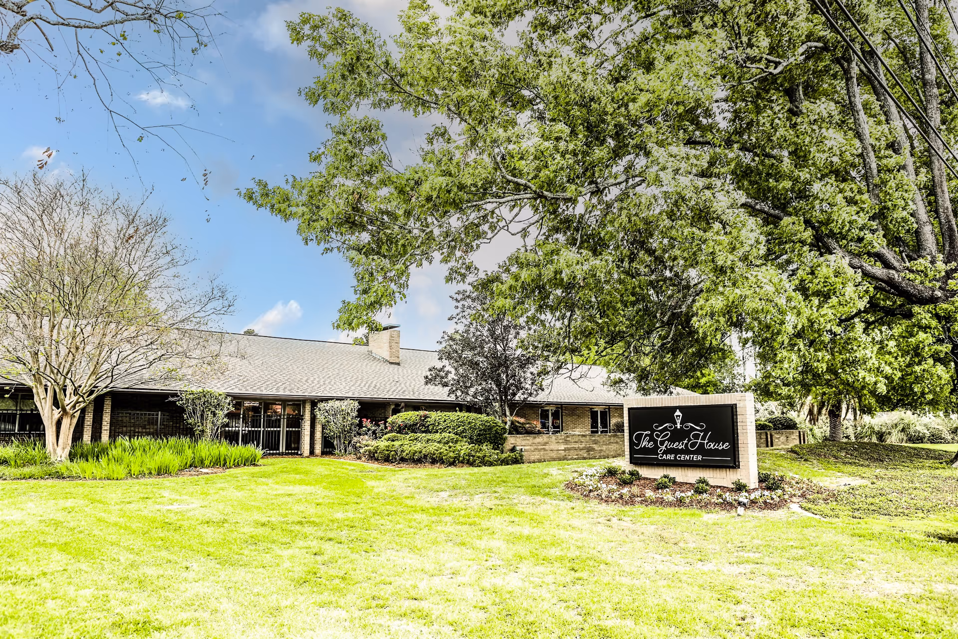The Guest House care center building with sign, green lawn, and large front trees.