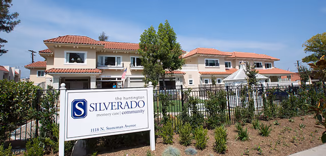 Front exterior of Silverado The Huntington memory care community, a two-story beige building with red tile roof, landscaping, fence, and a large sign.