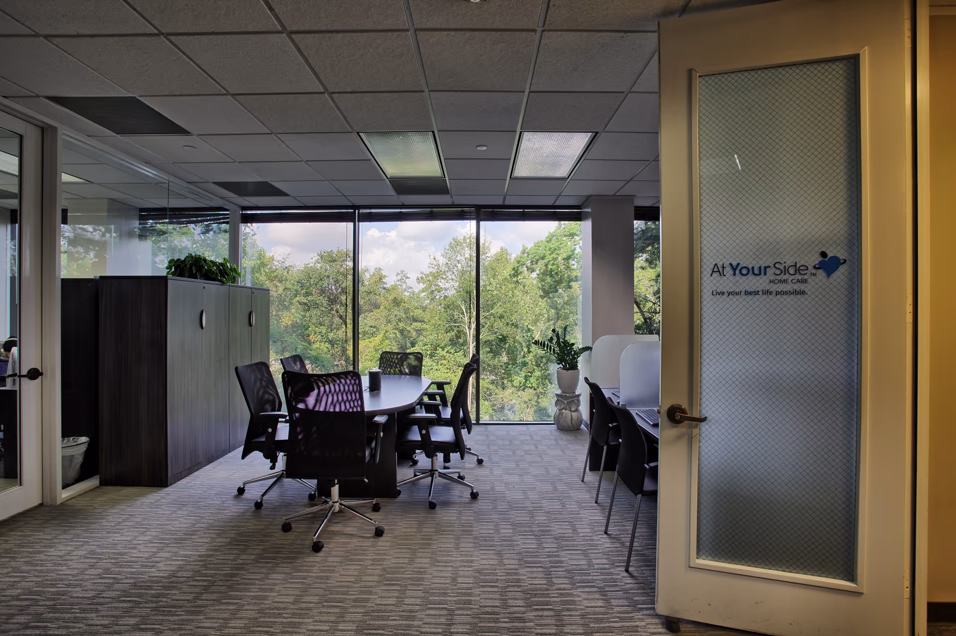 Indoor conference room with a long table, chairs, and large windows overlooking trees.