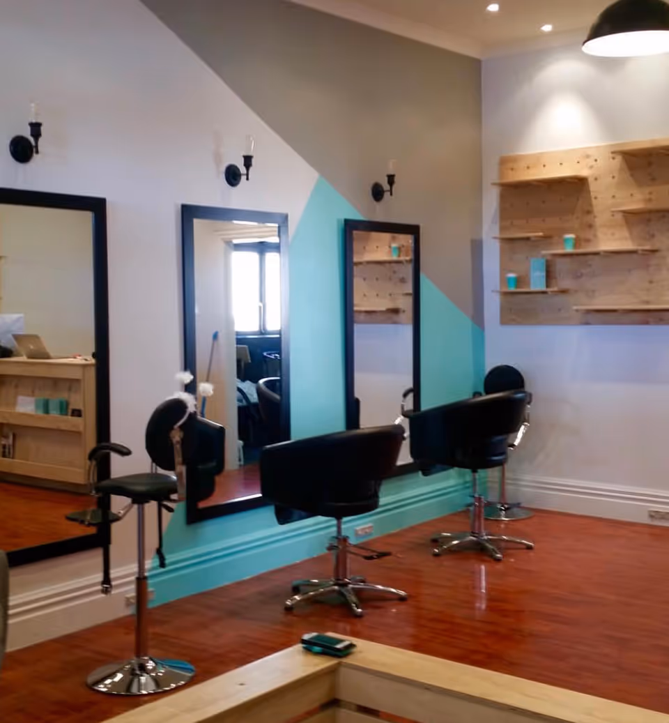 Modern hair salon interior with black chairs, large mirrors, turquoise trim, and wooden shelves.
