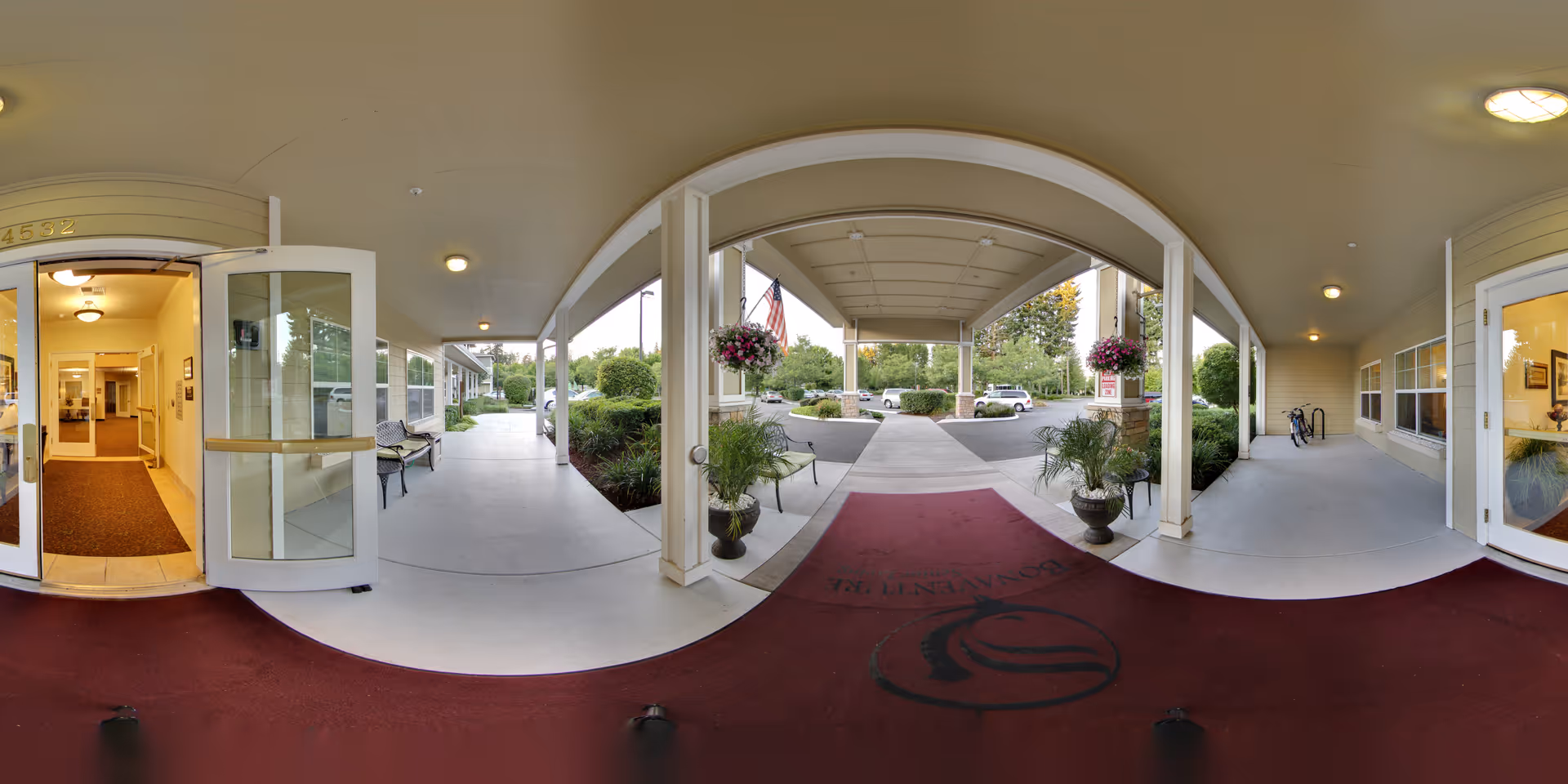 Covered entrance area of Woodland by Bonaventure senior living facility with open glass doors leading inside, benches along the walkway, potted plants, hanging flower baskets, and a parking area visible in the background.