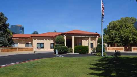 Single-story beige care facility with a red tile roof, trimmed shrubs, a flagpole, and a green lawn under a clear blue sky.