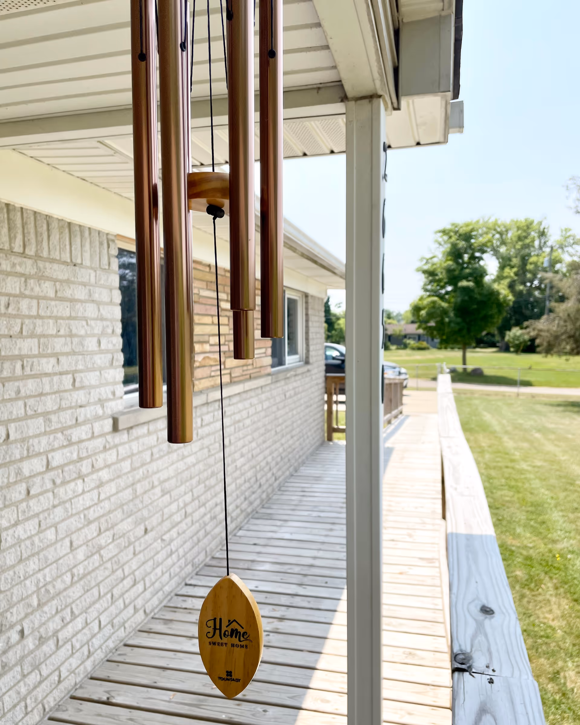 Wood wind chimes hang from a porch over a brick exterior with a grassy yard nearby.