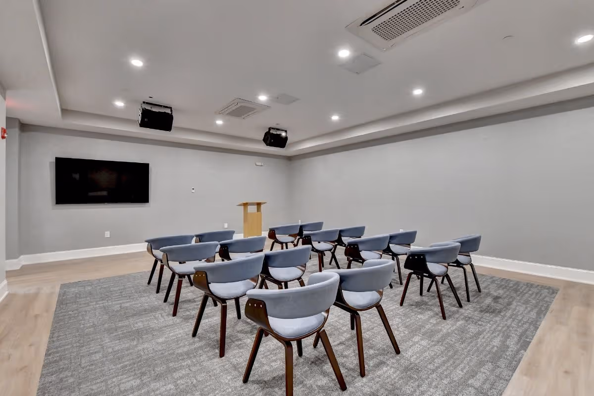 Interior conference room with podium, wall-mounted screen, gray carpet, and blue chairs.