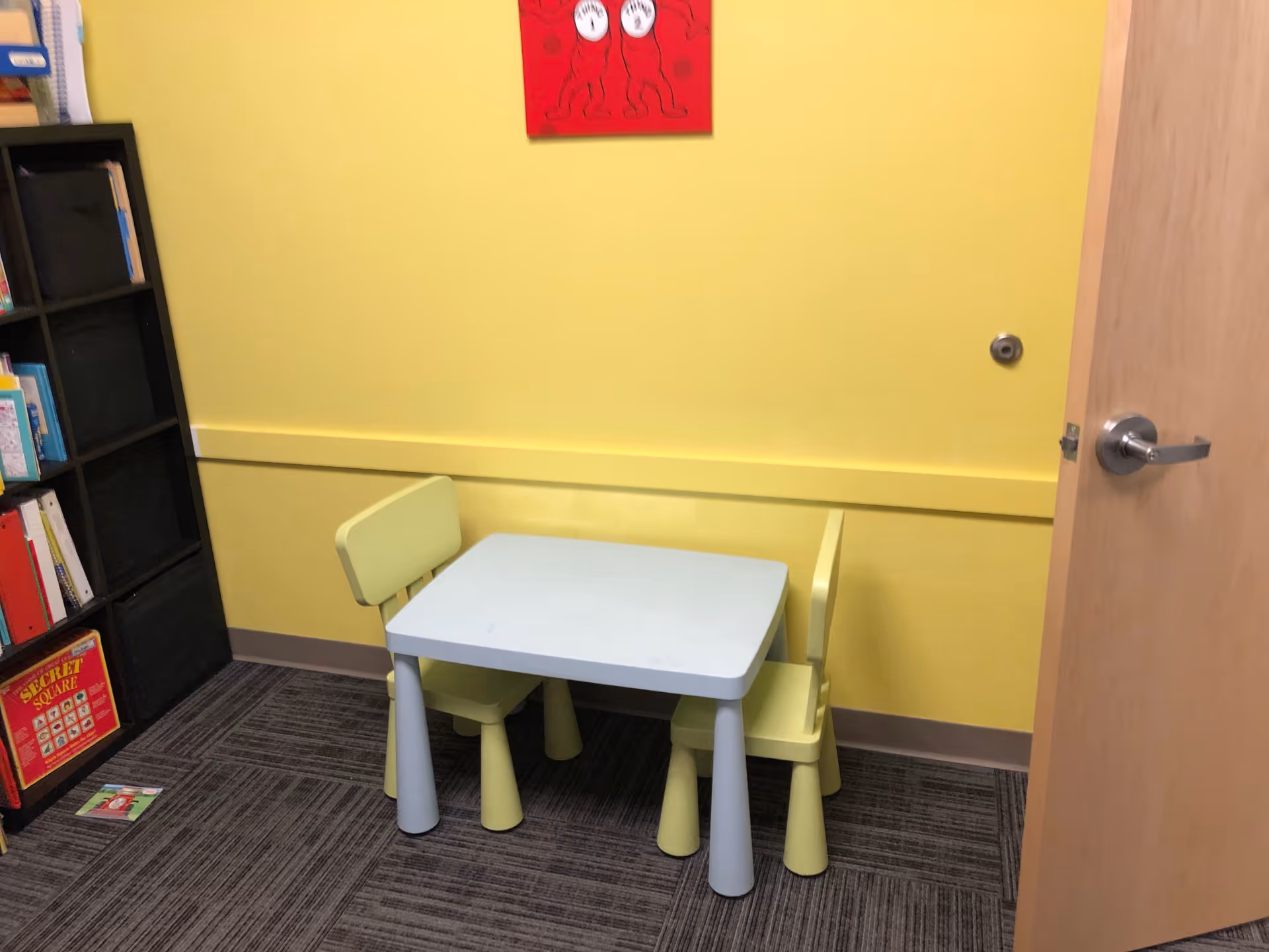 Yellow-walled room with a small blue table and pale green chairs beside a bookshelf.