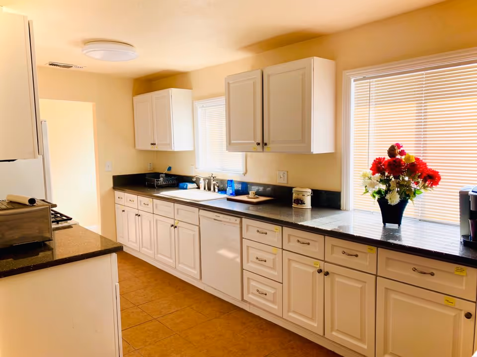 Bright kitchen with white cabinets and dark countertops, a sink under a window and a vase of red and white flowers on the counter.