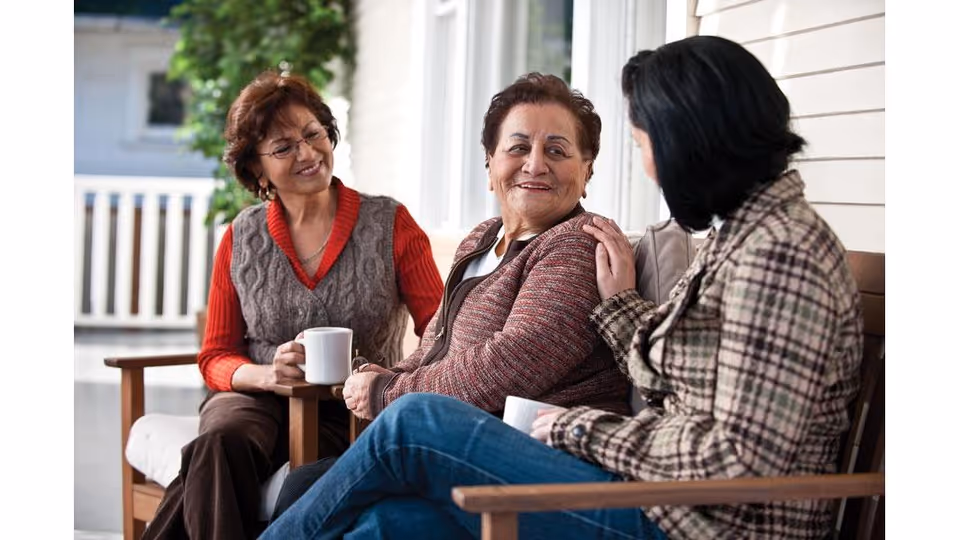 Three women sitting on a porch bench, engaged in conversation and holding white mugs. They appear relaxed and smiling, with a white railing and greenery visible in the background.