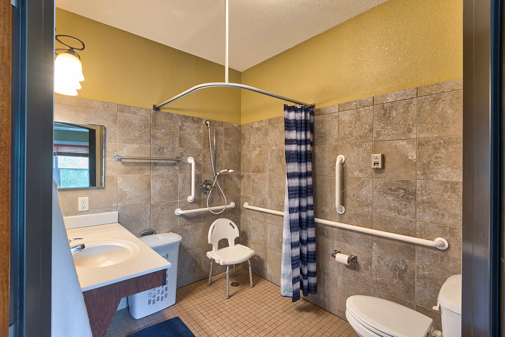 Accessible bathroom with shower chair, grab bars, and sink in tiled room.
