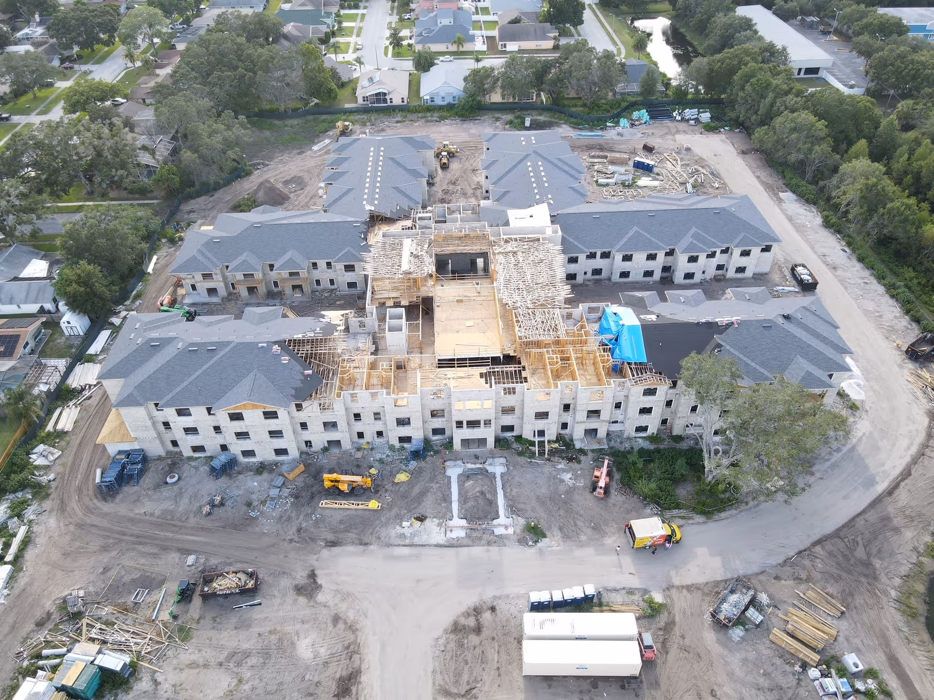Aerial view of a large senior living facility under construction, showing multiple connected buildings with roofs partially completed and wooden framing visible. Construction equipment and materials are scattered around the site, with residential houses and trees surrounding the area.