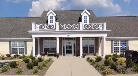 Front exterior view of a single-story assisted living community building with a gray shingled roof, two dormer windows, white columns supporting a porch, and a walkway leading to the entrance. The building is surrounded by landscaped bushes and plants, with a clear blue sky in the background.