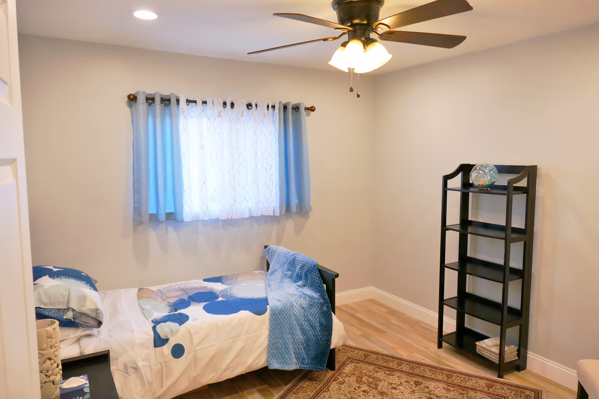 A small bedroom with a single bed, blue bedding, window, and tall shelf.