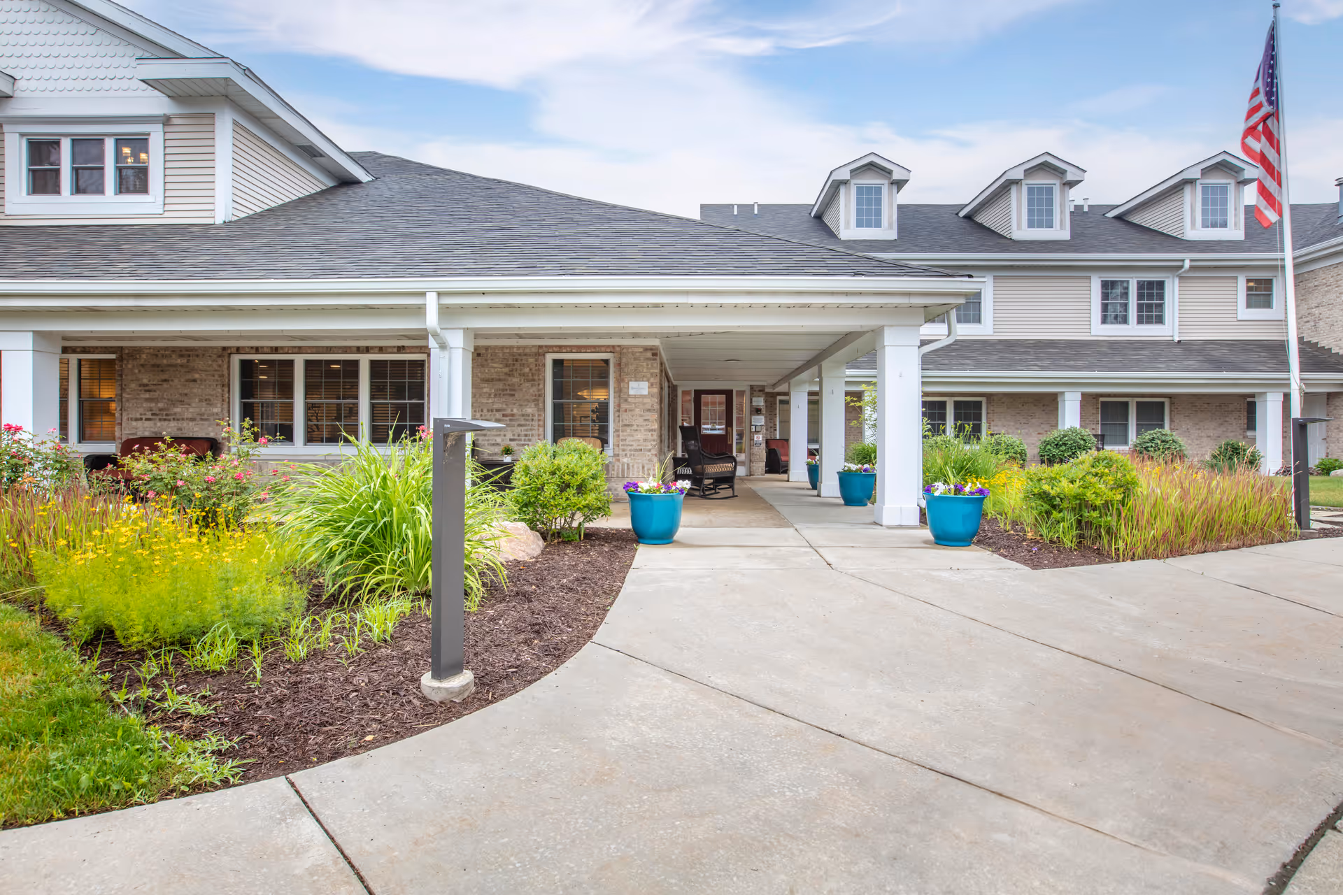 Front exterior view of Brookdale Meridian - Assisted Living facility showing a covered entrance with seating, landscaped garden beds with green plants and flowers, and an American flag on a flagpole to the right.
