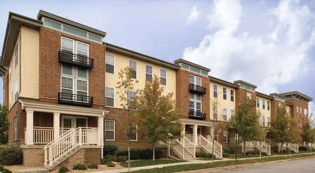 Exterior front view of a three-story brick and siding multi-unit residential building with stair entrances and small balconies.
