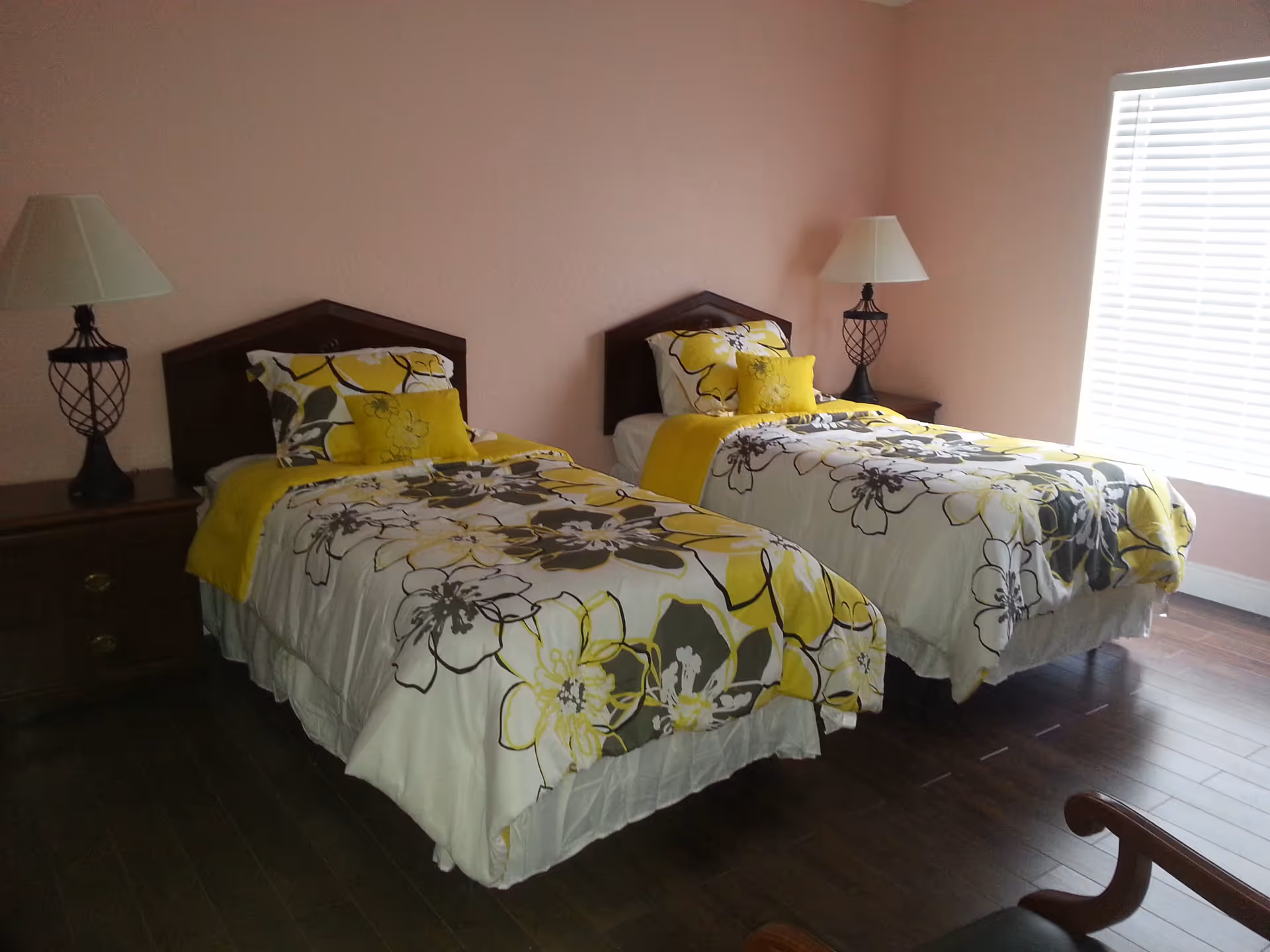 A bedroom with two twin beds featuring floral bedspreads in yellow, white, and gray. Each bed has a matching pillow and a small yellow decorative pillow. There are wooden nightstands on either side of the beds, each with a lamp. The room has pink walls, a window with white blinds, and dark wood flooring.