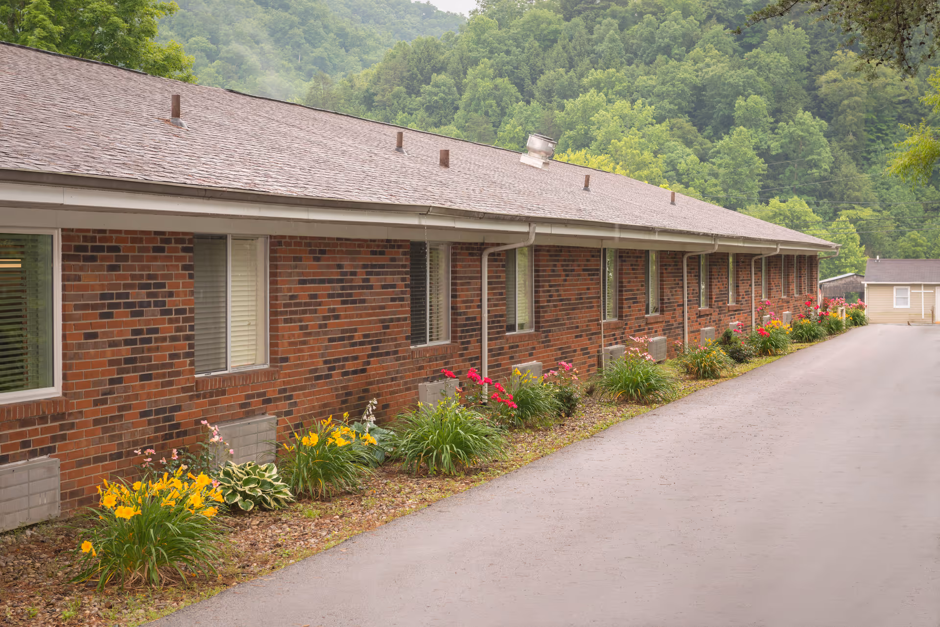 Long side view of a single-story brick building with multiple windows, a paved driveway, and flower beds with yellow and red flowers along the base. The building is surrounded by lush green trees and hills in the background.
