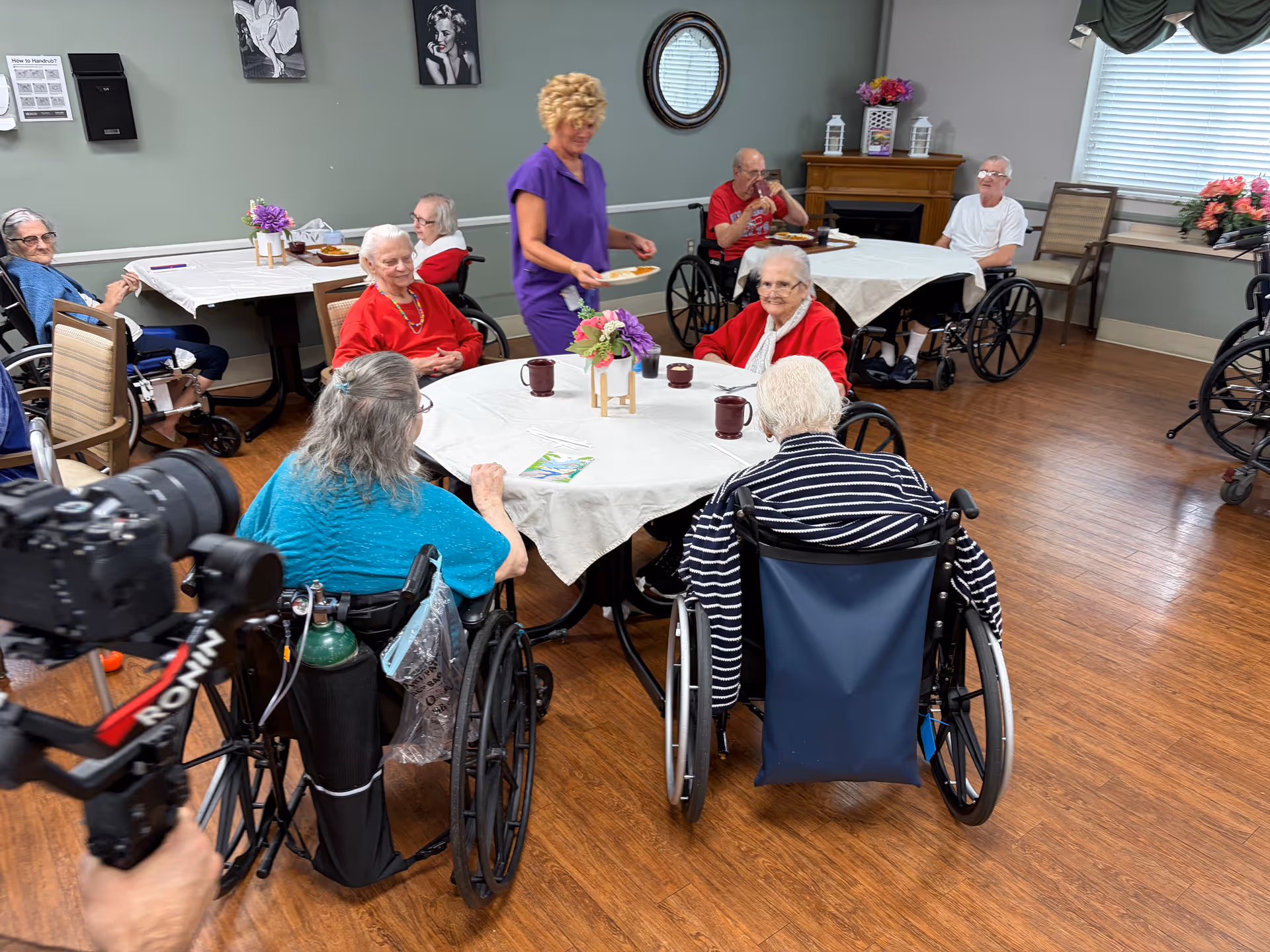 Caregiver serves plates to elderly residents in wheelchairs at round dining tables.