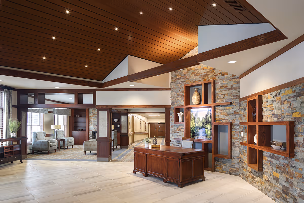 A spacious and well-lit assisted living facility common area with a high wooden ceiling featuring recessed lighting. The room has a stone accent wall with built-in wooden shelves displaying decorative items and a large framed picture of a forest and river. In the center, there is a wooden desk with a small plant and lantern on top. To the left, there is a seating area with upholstered armchairs and a side table near large windows letting in natural light. The floor is tiled in light colors, and the overall decor is warm and inviting with wood and stone elements.