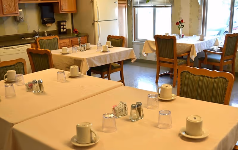 A dining room with several tables covered in beige tablecloths, each set with cups, glasses, and condiments. Wooden chairs with green cushioned seats surround the tables. The room has large windows letting in natural light and wooden kitchen cabinets along one wall.