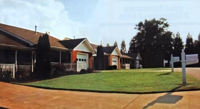 Exterior view of a single-story residential care facility with multiple attached units, each having a garage and a small front porch. The building is surrounded by a well-maintained lawn and some tall trees in the background. There are white mailbox posts near the driveway.