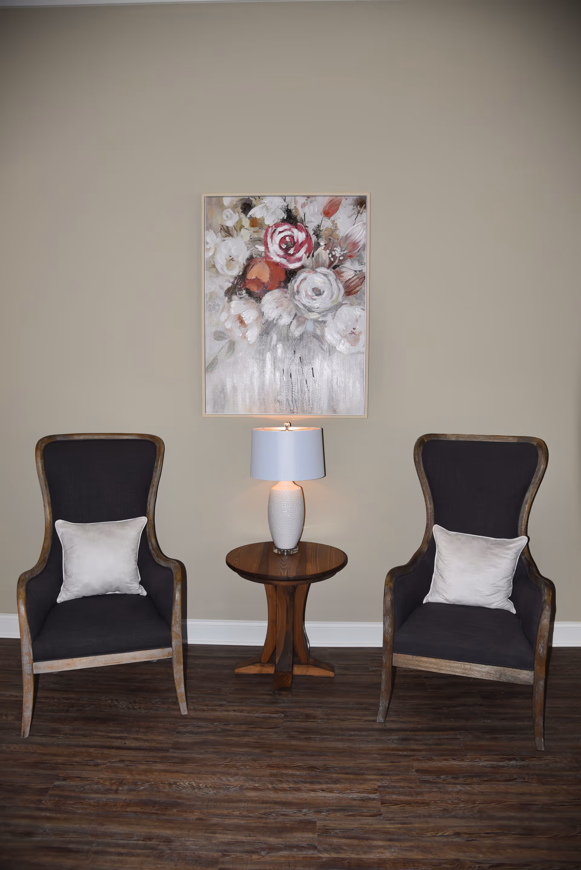 Two dark upholstered armchairs flank a wooden side table with a lamp beneath a floral painting on a beige wall.
