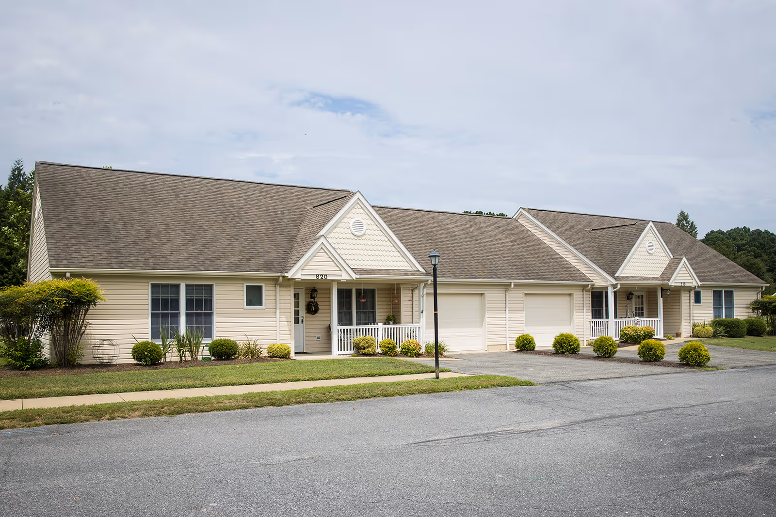 Exterior view of a single-story residential building with beige siding and brown shingled roof. The building features two units with small front porches, white railings, and attached garages. There are small bushes and plants along the front, a sidewalk, and a street in the foreground under a partly cloudy sky.