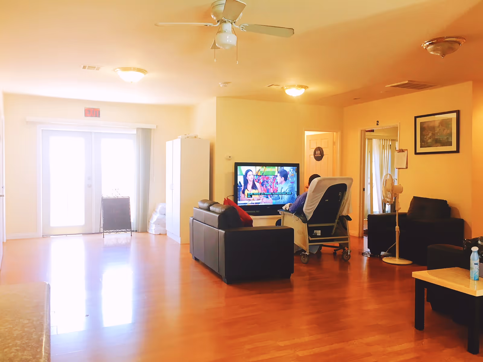 A bright senior living lounge with TV, sofa, recliner, cabinet, and sliding doors.