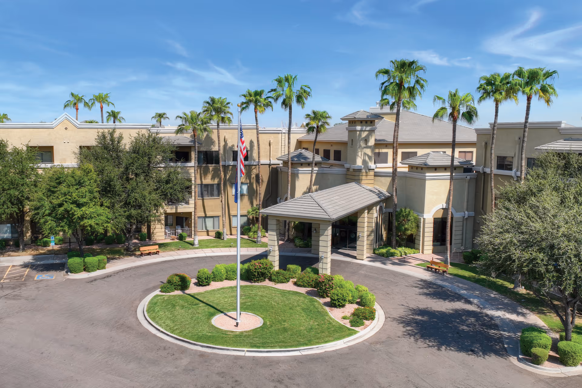 Front entrance of a senior living facility with a circular driveway, flagpole, and palm trees.