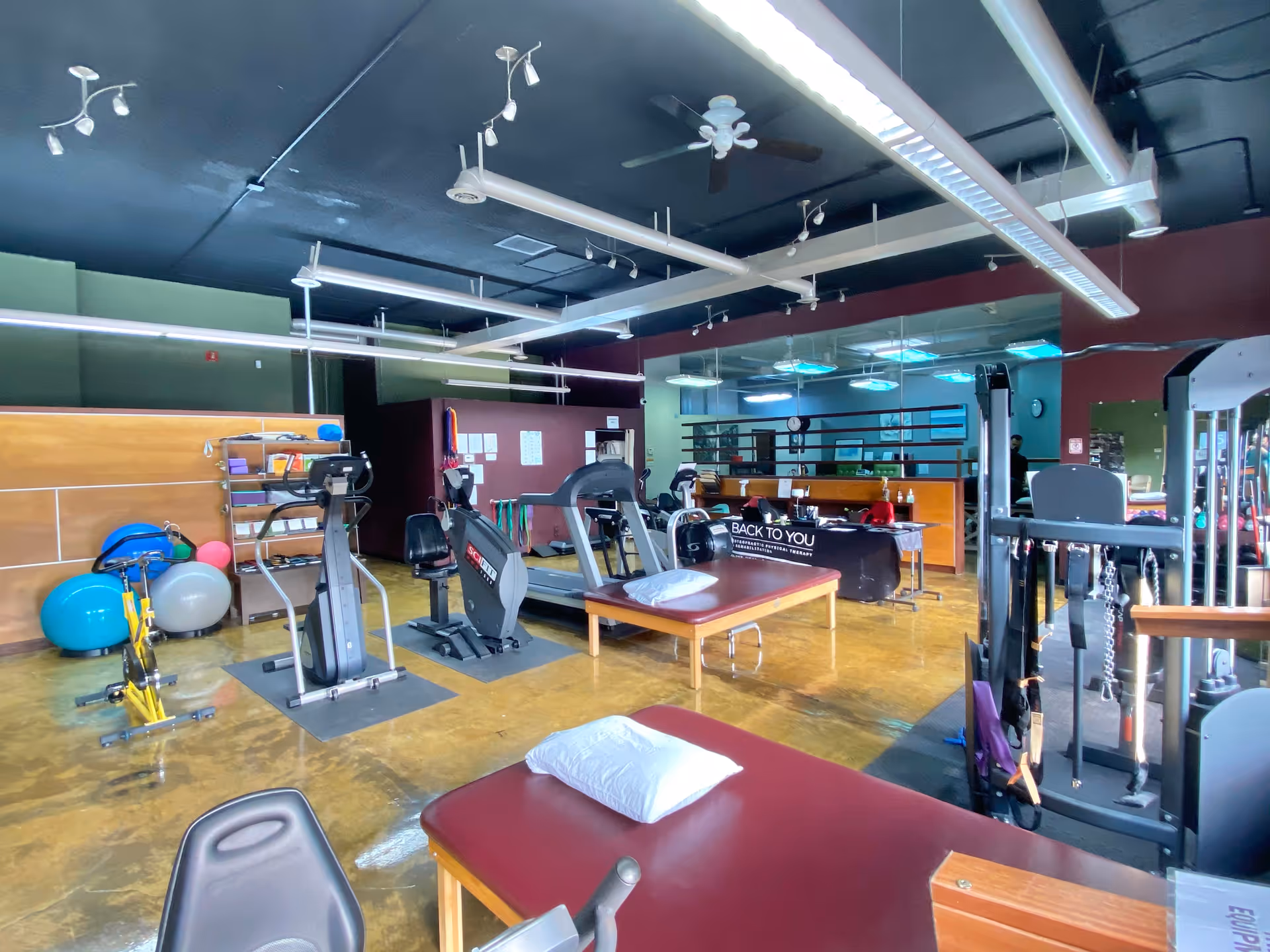Well-equipped rehab gym with cardio equipment, therapy tables, and a reception desk.