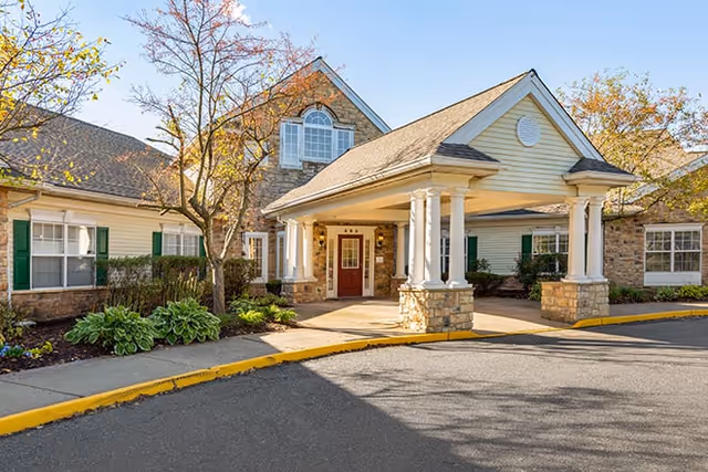 Front entrance of a residential senior living facility with a covered portico supported by columns and landscaped grounds.