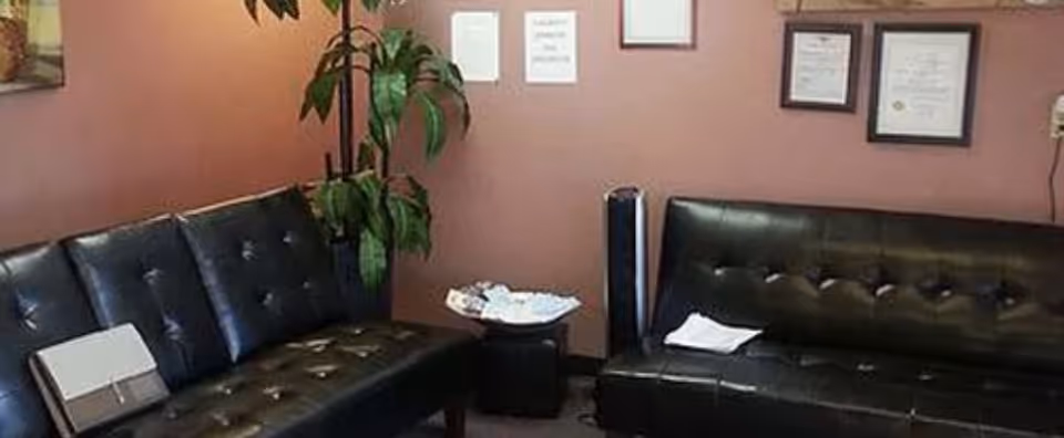 Waiting area with black leather sofas, plant, and framed certificates on pink wall.