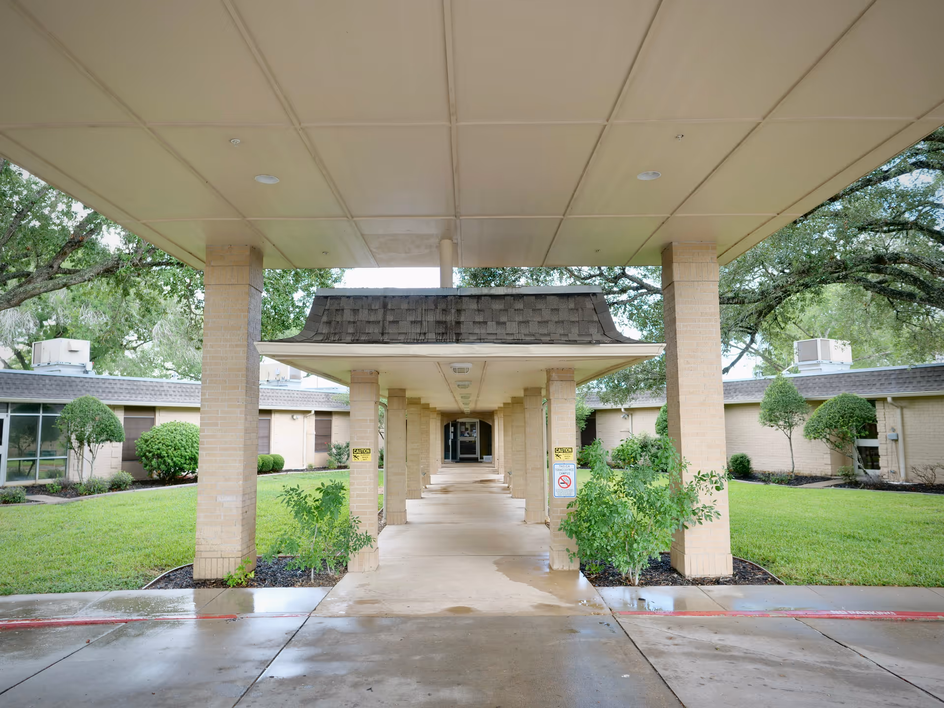 Covered entrance walkway with brick columns leading to the front doors of a low single-story nursing facility, flanked by lawns and trimmed shrubs.