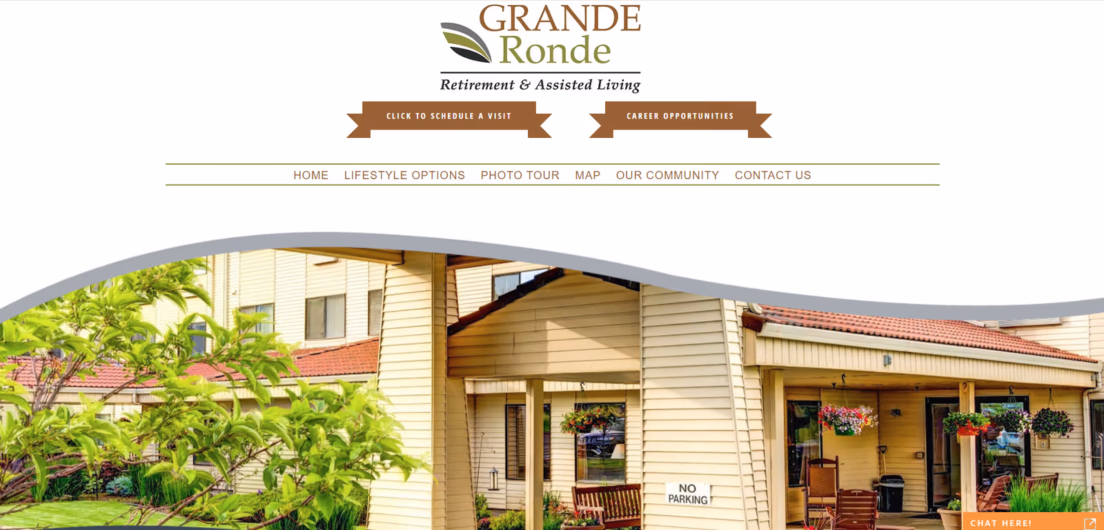 Exterior view of Grande Ronde Retirement & Assisted Living facility showing a beige building with a red roof, hanging flower baskets, outdoor seating, and greenery in the foreground.