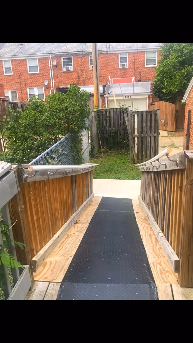 Outdoor wooden ramp with handrails leads to a fenced backyard and brick buildings.
