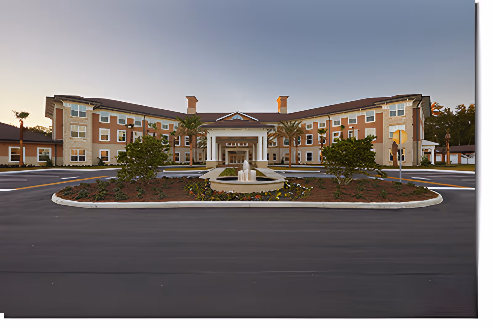 Front exterior view of a large, multi-story senior living facility building with a central entrance featuring white columns and a covered driveway. There is a landscaped roundabout with a small fountain and greenery in front of the building. The sky is clear and the area is well-maintained.