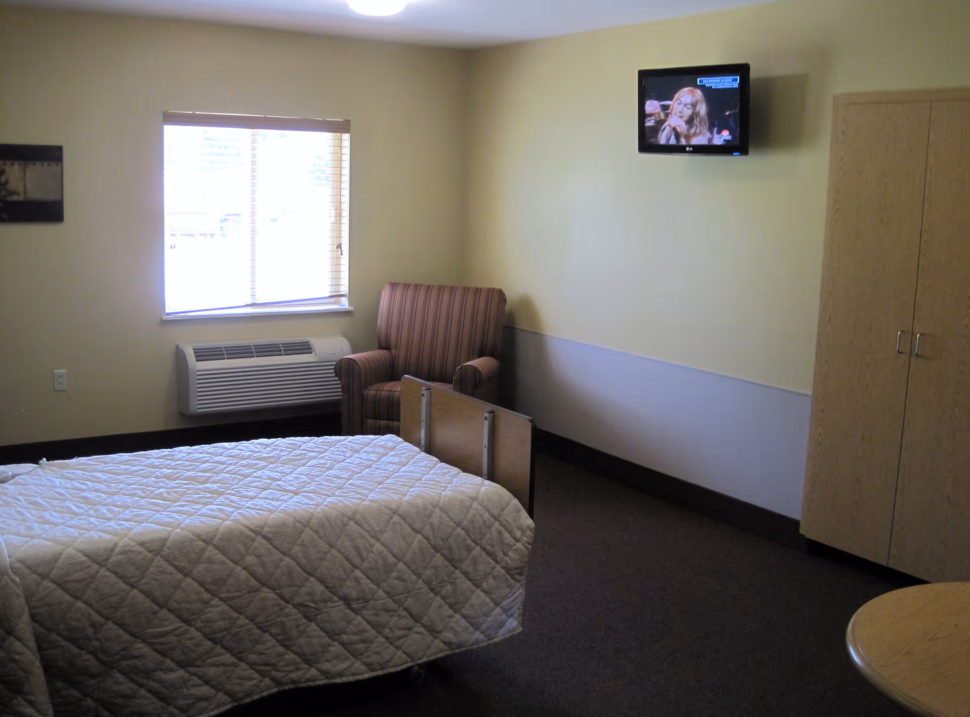 A simple bedroom with a single bed covered in a white quilted blanket, a striped armchair near a window with blinds, a wall-mounted TV showing a woman singing, a wooden wardrobe, and a small round table in the corner.