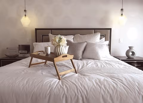 Neatly made bed with decorative pillows and a wooden breakfast tray holding a vase of flowers, flanked by pendant lights and bedside tables.