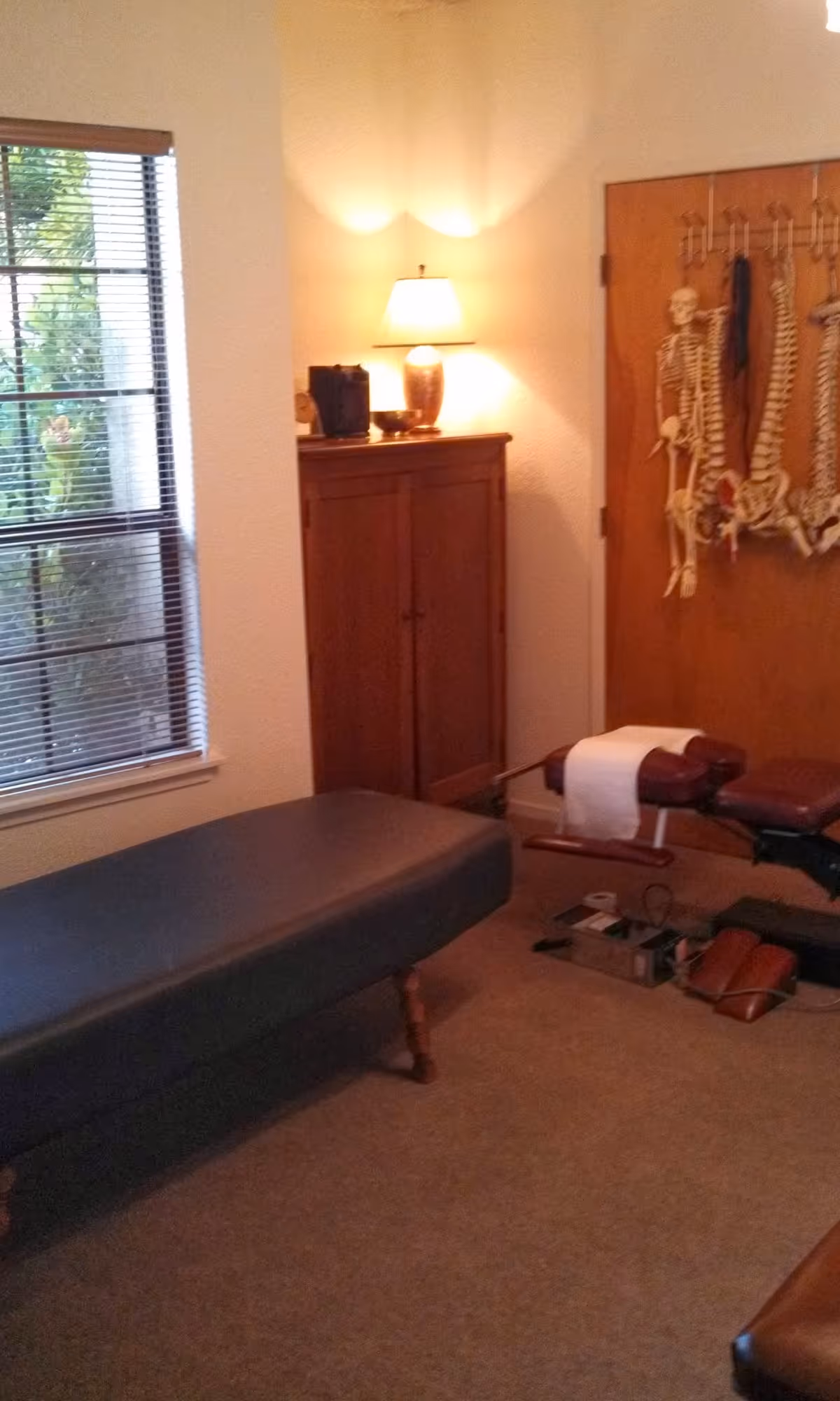 A small therapy room with a treatment table, cabinet, skeleton poster, and warm lamp.