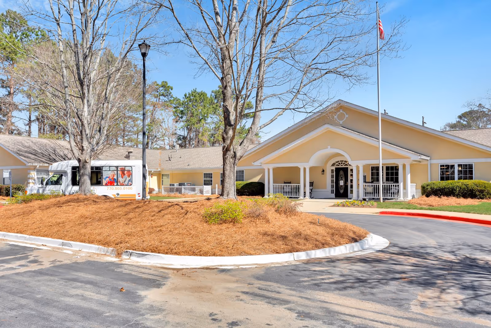 Exterior view of Peachtree Creek Memory Care assisted living facility in Georgia, showing a single-story building with a covered entrance, an American flag on a flagpole, a circular driveway, and a white shuttle bus parked near the building. Trees and landscaping are visible around the property under a clear blue sky.