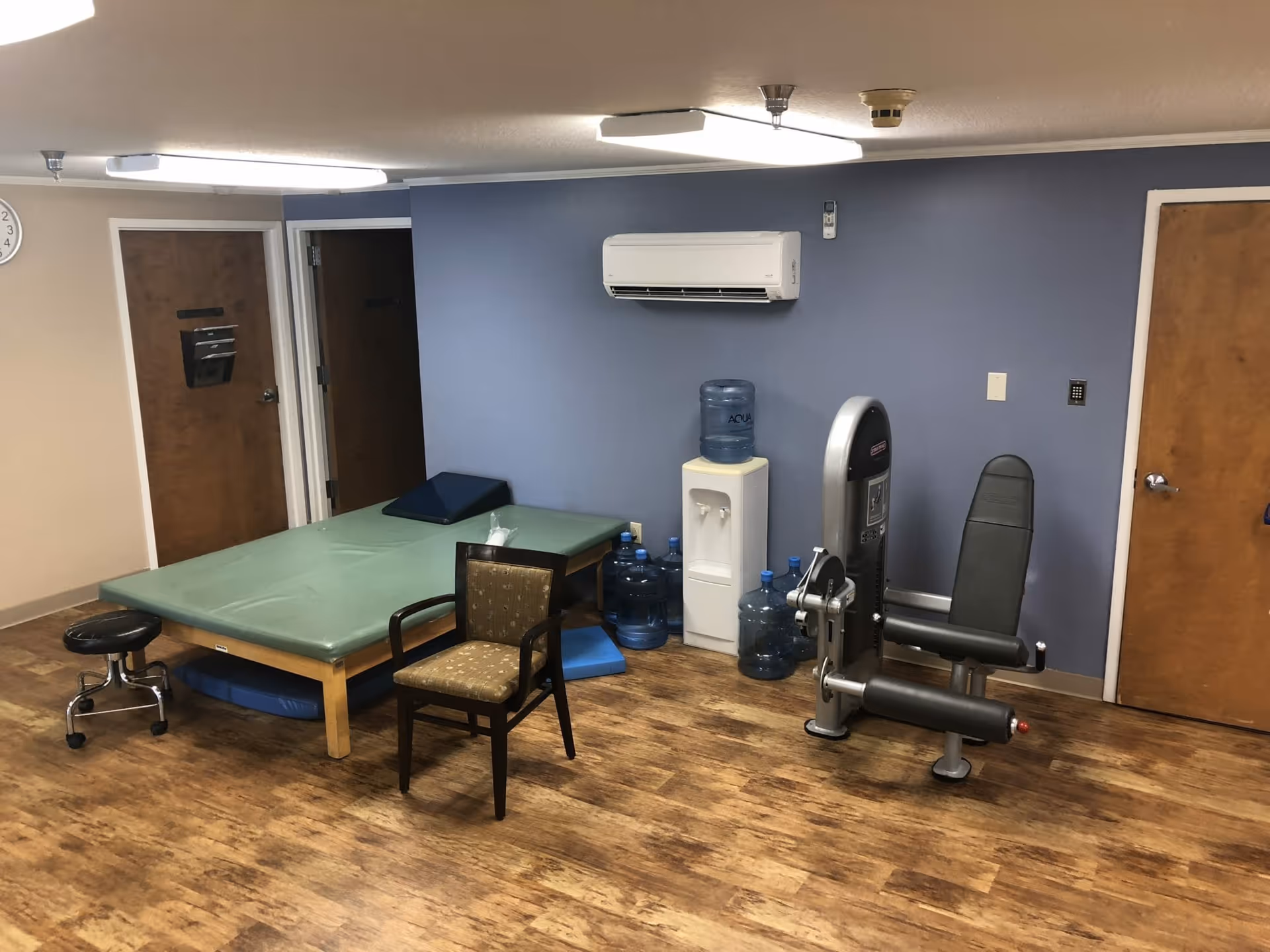 A rehabilitation room with a padded treatment table, a chair, a stool on wheels, a leg exercise machine, a water cooler with several water jugs, and a wall-mounted air conditioning unit. The room has wood flooring, blue and beige walls, and two closed wooden doors.