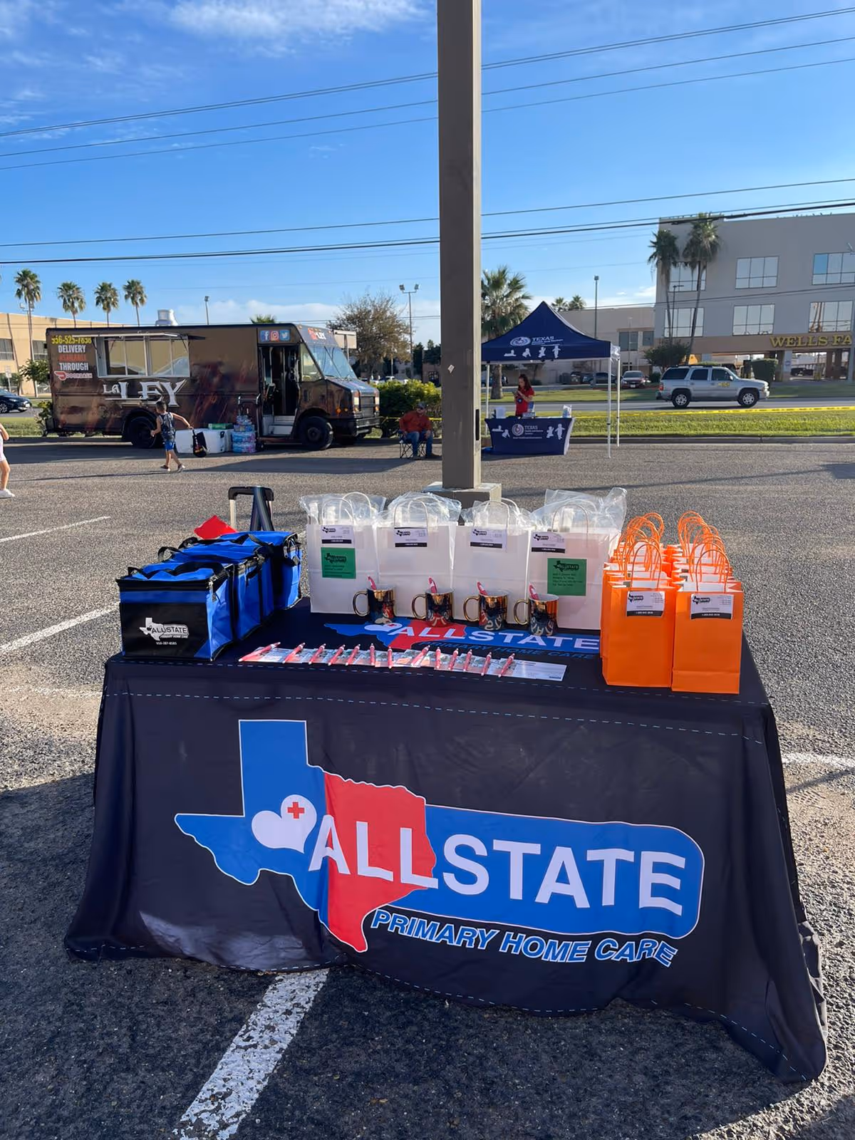 Outdoor event table with Allstate Primary Home Care branding and giveaway bags in parking lot