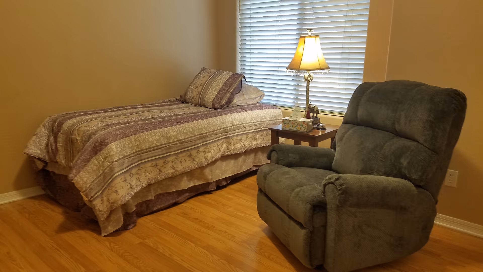 A small bedroom with a single bed covered in a patterned bedspread and a matching pillow. Next to the bed is a wooden side table with a decorative lamp, a tissue box, and a small elephant figurine. A large, comfortable recliner chair is positioned near the side table. The room has wooden flooring and a window with closed blinds behind the side table and lamp.